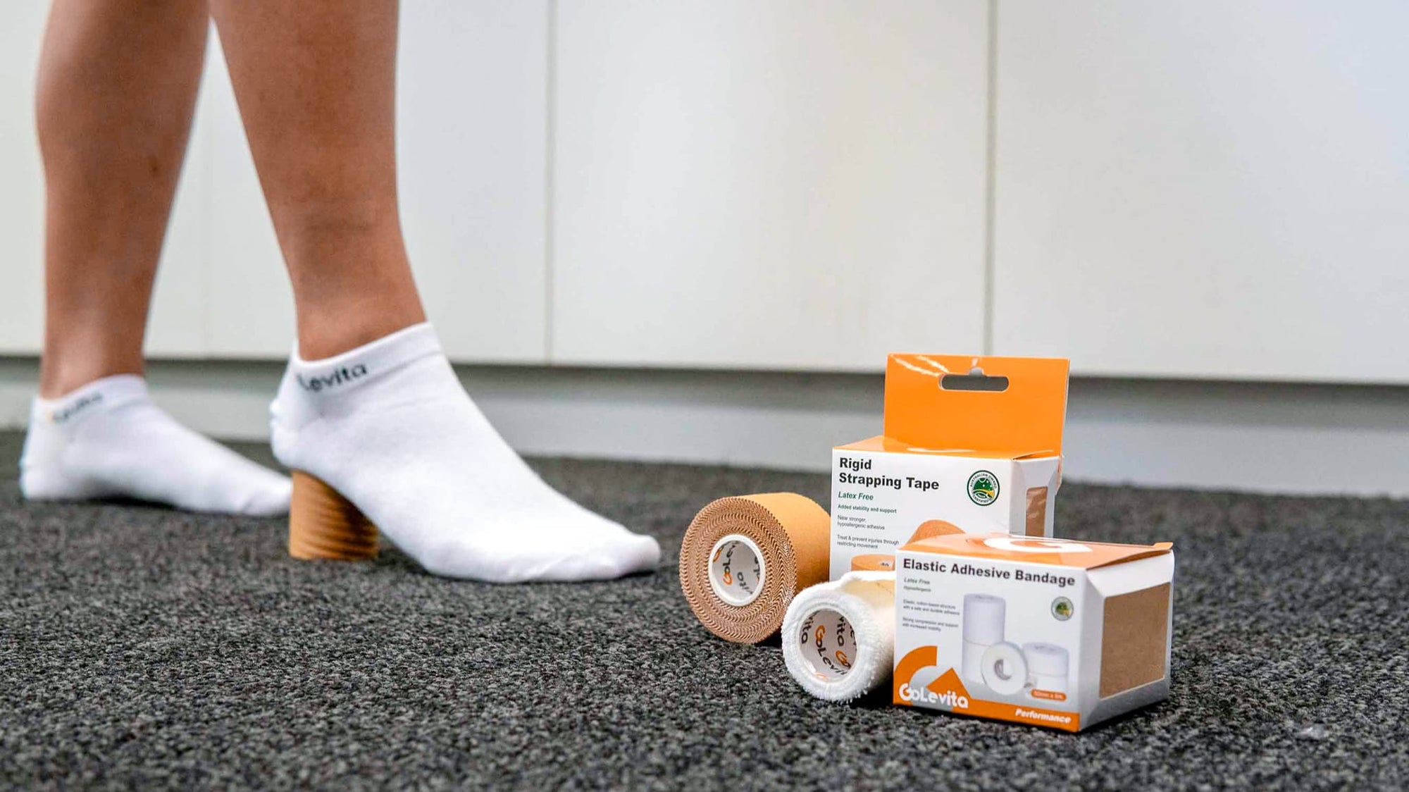 Person wearing white socks with a GoLevita logo, next to GoLevita medical tape and bandage packaging on a dark surface in physiotherapy clinic for educational content on strapping tape Australian made